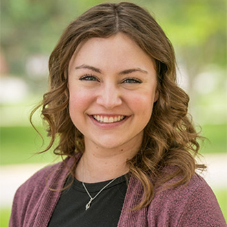 A young woman with wavy brown hair, wearing a maroon sweater, smiles outdoors on a Minnesota campus, representing student engagement.
