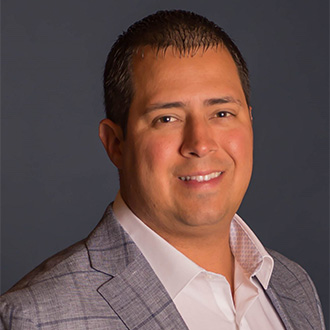 A man with short dark hair, wearing a light gray plaid suit, smiles; he represents student engagement at Bethany Lutheran College.