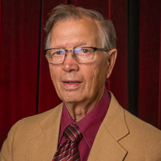 A man wearing glasses, a tan jacket, a red shirt, and a striped tie stands in front of a red curtain at Bethany Lutheran College.