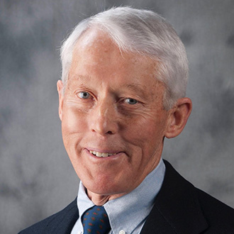 A man with short white hair, wearing a suit, blue shirt, and patterned tie, smiles at the camera at Bethany Lutheran College.