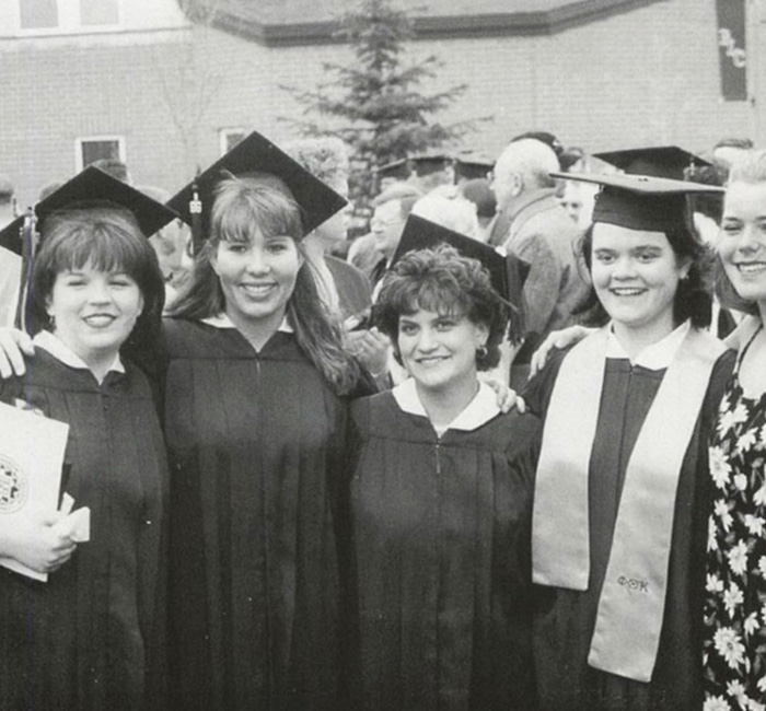 Five Bethany Lutheran College students in graduation gowns and caps smile together outdoors for a group photo.