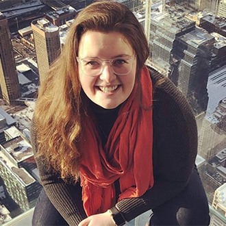 Smiling woman with long hair and glasses, wearing a red scarf, poses above a Minnesota cityscape with tall buildings below.