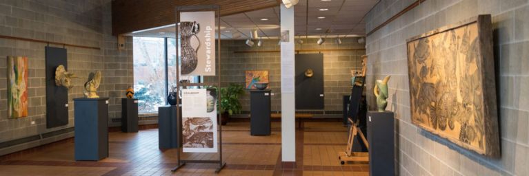 Art gallery with sculptures displayed on pedestals, paintings hung on the walls, and a large central sign that reads Stewardship.