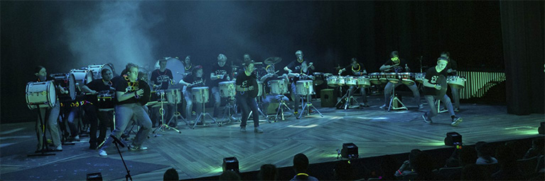 A group of drummers from Bethany Lutheran College perform energetically on stage, engaging the student audience.
