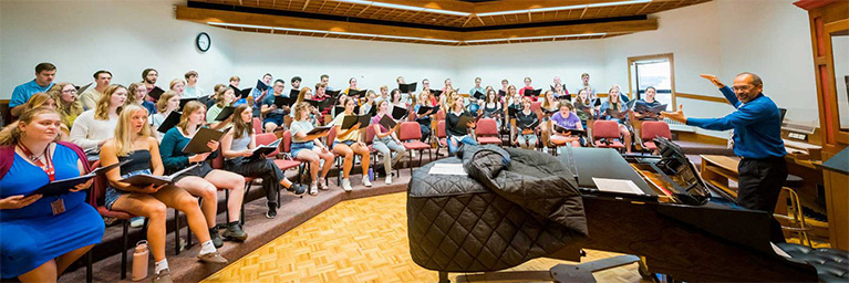 A large choir rehearses in a bright room, with a conductor leading the group near a grand piano.