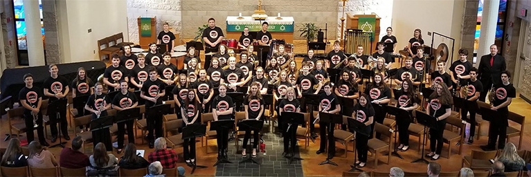 A large student band wearing matching shirts poses with their instruments inside a church, demonstrating campus community spirit.