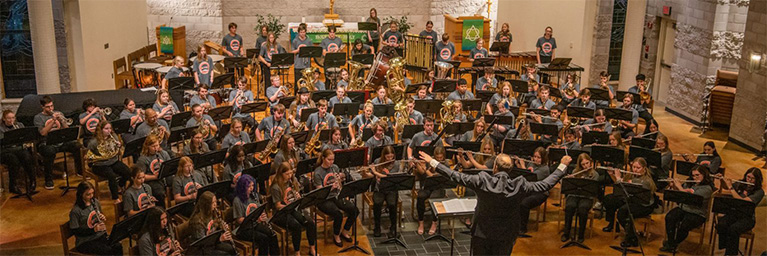 A large ensemble of musicians from Bethany Lutheran College performs on a church stage, directed by a conductor.