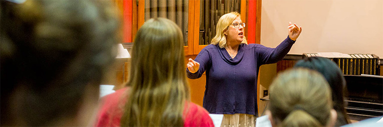 A woman conducts a choir of students in Mankato, with a piano and organ pipes visible in the background.