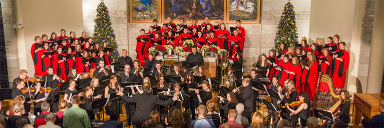 A choir in red robes sings behind an orchestra on stage, while Mankato’s campus community gathers near decorated Christmas trees.
