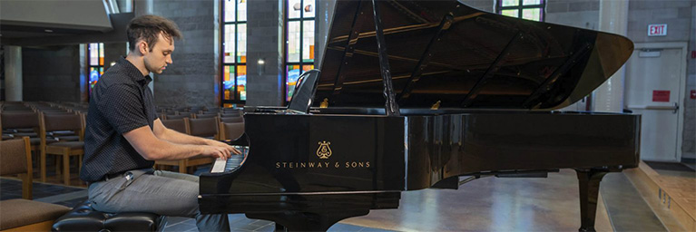 A man plays a Steinway & Sons grand piano in a large room at Bethany Lutheran College, featuring tall stained glass windows.