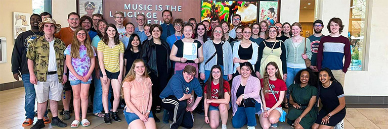 A large group of smiling young adults in Minnesota poses indoors in front of a sign about student engagement in music and art.