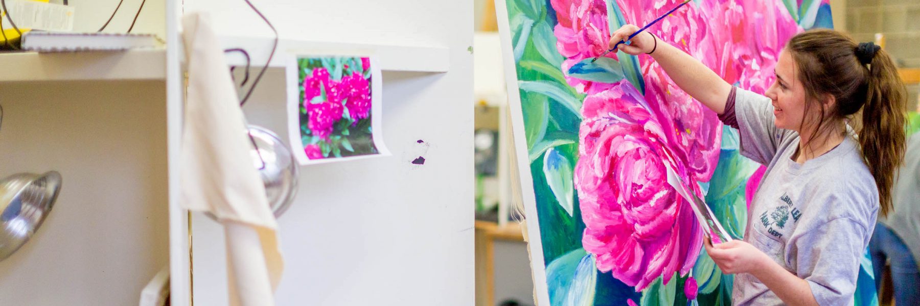 A Bethany student paints large pink flowers on a canvas, using a printed photo taped to a nearby shelf as reference.