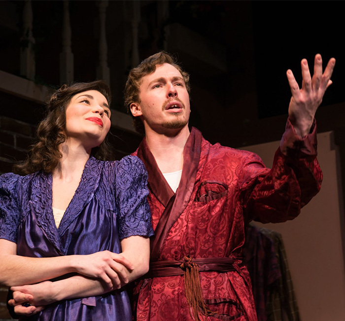 A student in a purple dress stands on stage beside a man in a red robe who is gesturing, during an event at Bethany Lutheran College.