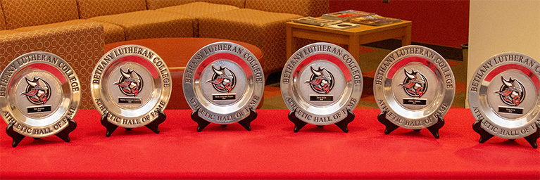 Six plaque awards featuring mascot logos are arranged on a red tablecloth, recognizing achievements within the Mankato campus community.