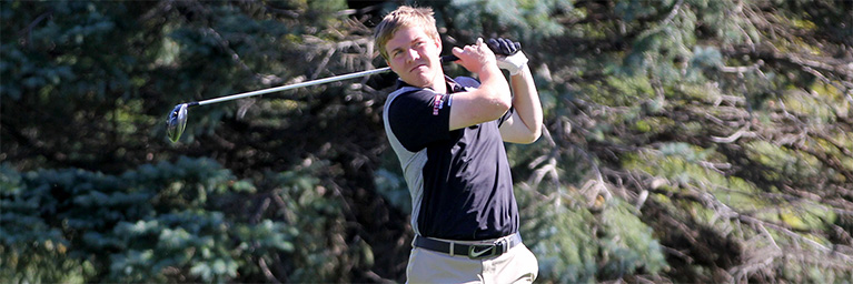 A golfer wearing a black shirt and beige pants swings a club outdoors on the Mankato campus, with trees visible in the background.