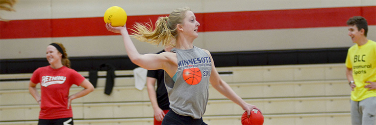 A young woman throws a yellow dodgeball in a Bethany Lutheran College gym while students watch and smile in the background.