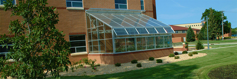 A small glass-walled greenhouse is attached to a brick building on the Mankato campus, surrounded by green grass.