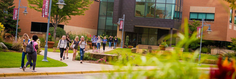 Students walk along a sidewalk at Bethany Lutheran College, with brick buildings and greenery visible in the background.