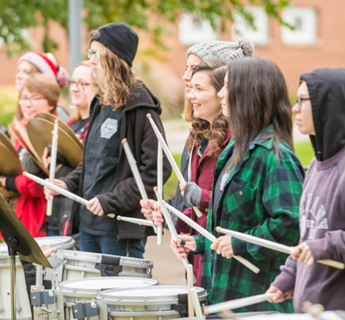 Bethany Lutheran College students play snare drums and cymbals outdoors, smiling in casual attire.