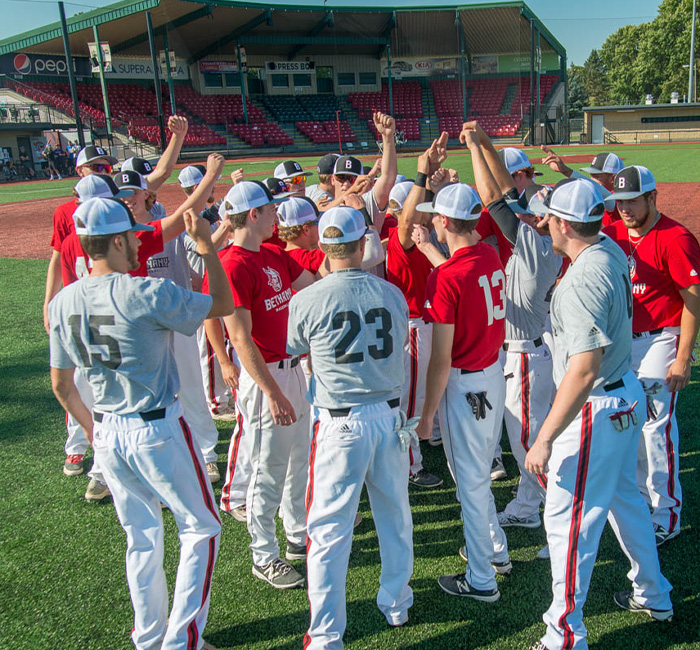 Bethany Lutheran College students from Mankato, wearing red and gray shirts, huddle on the field with hands raised before a baseball game.