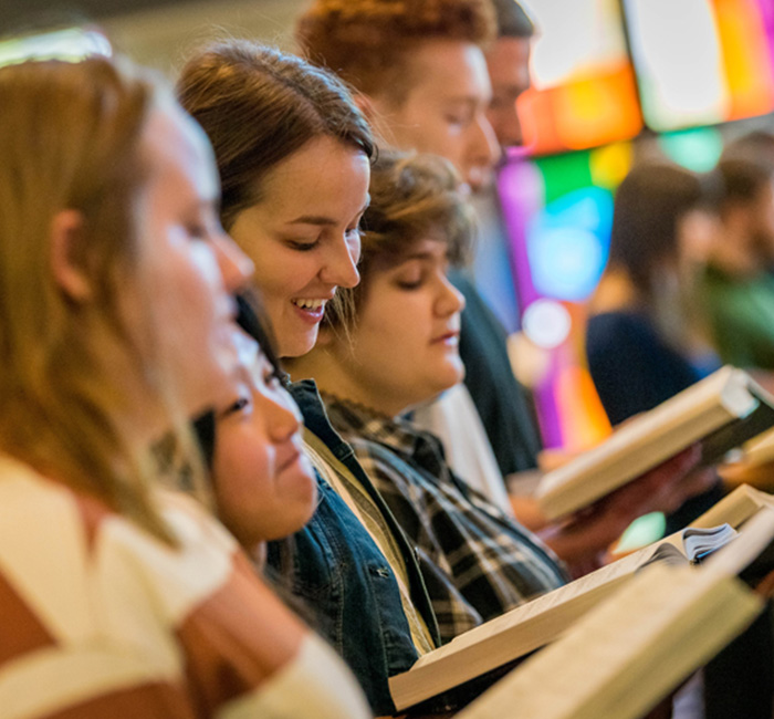 Students at Bethany Lutheran College sing together, holding books before colorful stained-glass windows in Minnesota.