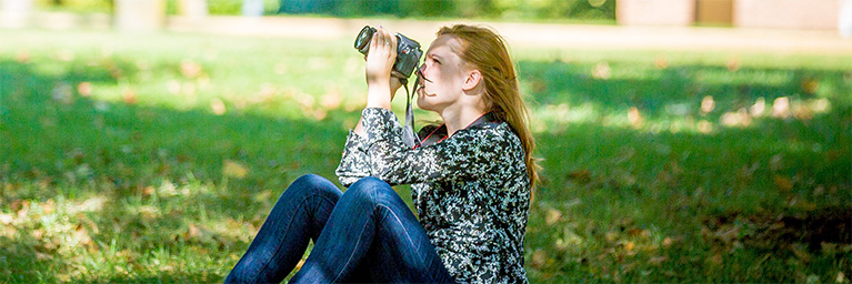 A woman sits on the grass at a Minnesota campus, holding a camera and looking up, surrounded by sunlight and green trees.