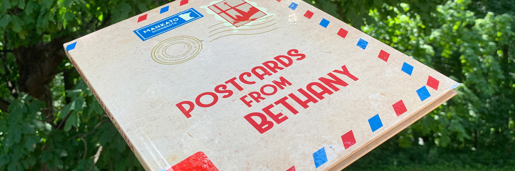 A postcard-style envelope labeled Postcards from Bethany is held up outdoors in Mankato, Minnesota, with green trees visible in the background.