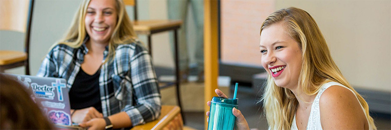 Two young women smile and talk; one holds a drink cup while the other types on a laptop, connecting in their campus community.