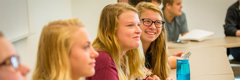 Students seated at desks in a Minnesota classroom, smiling and engaged, with notebooks and a water bottle on the table.
