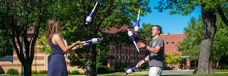 Two people juggle clubs outdoors on the Bethany Lutheran College campus, with trees and a brick building in the background.