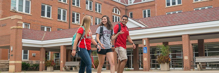 Three college students with backpacks stand outside a brick building at Bethany Lutheran College, smiling and talking together.