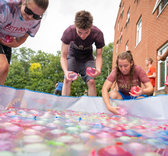 Three Bethany Lutheran College students lean over a pool filled with colorful water balloons outside a brick building in Mankato.