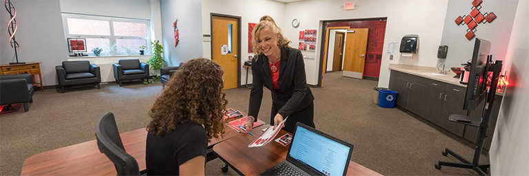 Two women interact at a desk in a modern Minnesota office, with chairs, a computer, and wall decorations visible in the background.