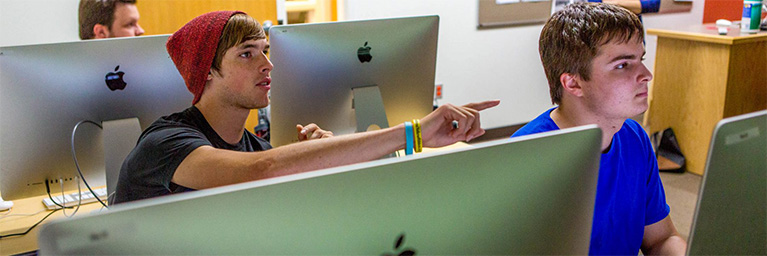 Two students sit at iMac computers on the Bethany Lutheran College campus; one points at the screen while the other appears focused.