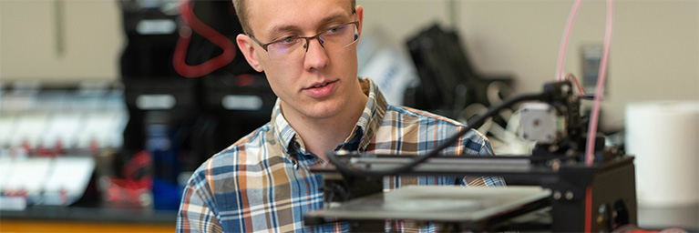 A person wearing glasses examines a 3D printer in a laboratory, demonstrating student engagement at Bethany Lutheran College.