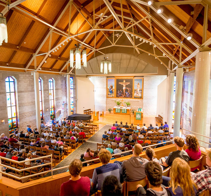 People seated in the Bethany Lutheran College Chapel with stained glass windows, listening to a speaker at the altar.