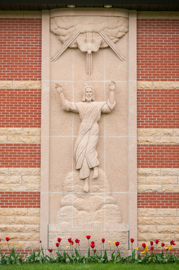 Stone relief depicting Jesus with raised arms ascending above tulips, set on a brick and stone building facade.