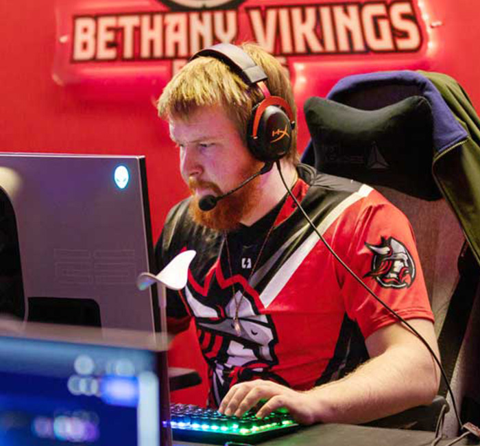 A gamer wearing a headset plays at a computer, with a Bethany Vikings sign visible in the background.