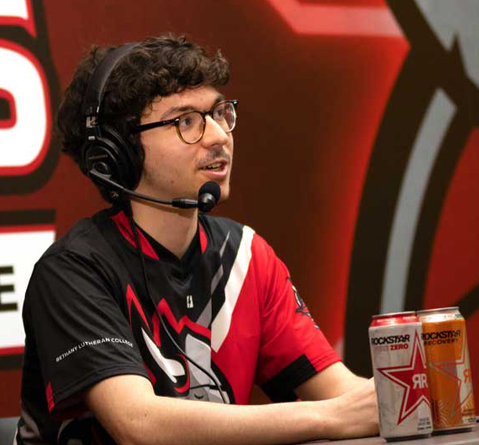 A person wearing a headset speaks at a gaming event table, with two Rockstar cans placed in front of them.