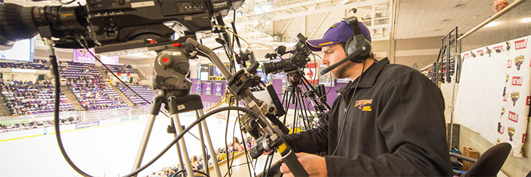 A cameraman wearing a headset films a Minnesota hockey game from the stands, documenting student engagement in the arena.
