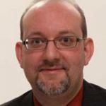 A man with glasses, short hair, and a goatee, dressed in a dark suit jacket and red shirt at Bethany Lutheran College, Minnesota.