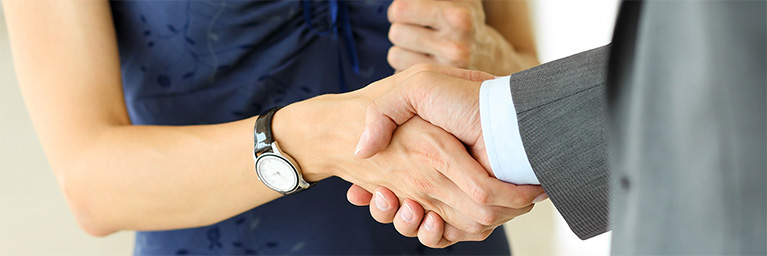 Close-up of a handshake between two individuals, one wearing a suit and the other wearing a dress, symbolizing partnership within the campus community.