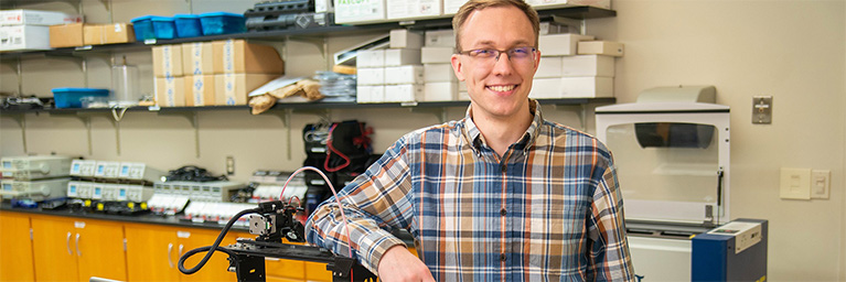 A smiling person wearing glasses stands next to lab equipment, representing student engagement in the Bethany Lutheran College campus community.