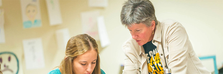 A teacher assists a student with schoolwork in a classroom at Bethany Lutheran College, with drawings displayed on the walls.