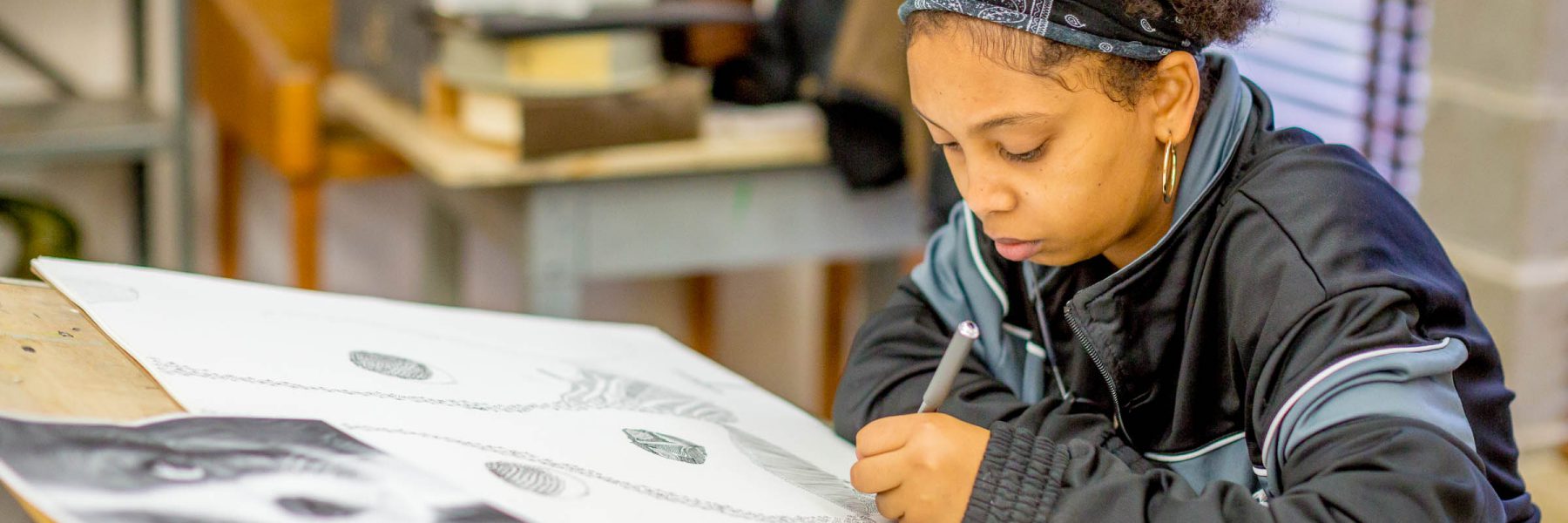 A young Bethany student sits at a table in an art studio, focused on drawing detailed artwork with a pencil.