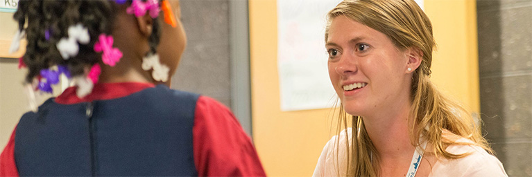 A woman smiles and talks with a young girl wearing colorful hair clips in a busy campus hallway.