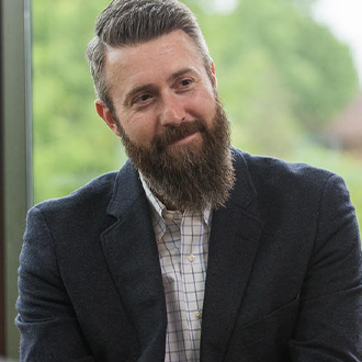Ben Kohls, Director of Graduate Studies in Clinical Mental Health Counseling for the Graduate Programs Department and Online Instructor for the Academic Affairs Department, with greenery in the background.