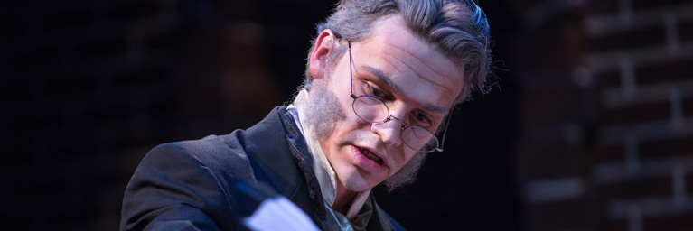 A student at Bethany Lutheran College, dressed in 19th-century costume and glasses, reads a paper under dramatic lighting against a brick wall in Mankato.