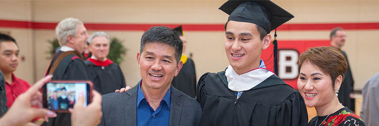 A graduate in a cap and gown poses with two smiling family members at Bethany Lutheran College in Mankato.
