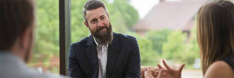 A bearded man in a suit listens attentively to two people talking in a bright room at Bethany Lutheran College.
